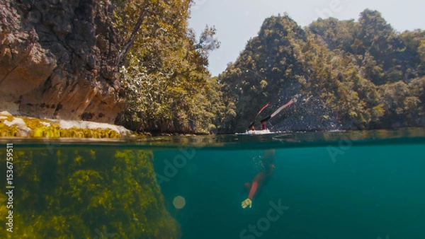 Obraz Freediver swims underwater in the closed lake. Splitted above and underwater view of the diver swimming in the lake surrounded by limestone mountains. West Papua, Misool, Indonesia