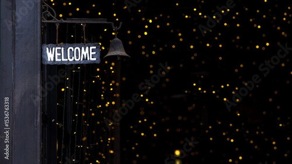 Obraz A, WELCOME, sign hangs on the dark black pole with a small bell. The background sparkles with numerous soft yellow lights, creating a warm and inviting atmosphere.