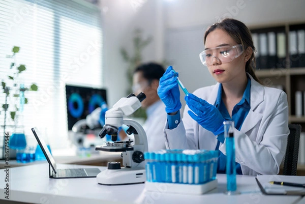 Fototapeta A woman in a lab coat is holding a pipette and looking at a microscope