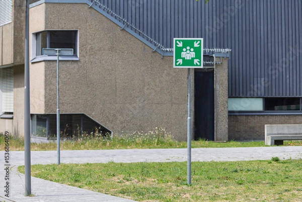 Obraz A bright green emergency assembly point sign stands prominently in front of a modern building, emphasizing safety.