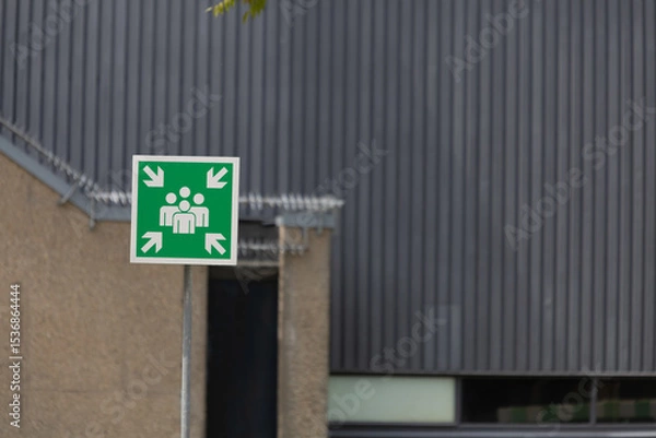 Obraz A prominent green emergency assembly point sign, set against a modern building facade, highlighting safety and preparedness.