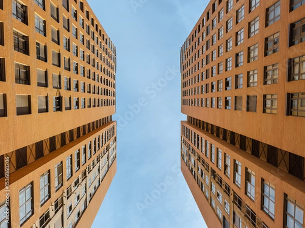 Obraz Striking upward view of two symmetrical modern high-rise buildings with orange facades, showcasing urban architecture, geometry, and city living under a clear blue sky.
