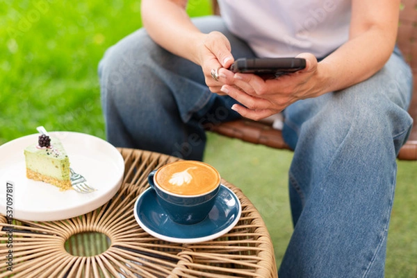 Fototapeta Close-up of a woman holding a cappuccino with latte art and a smartphone during a relaxing outdoors break featuring a plate with a dessert slice on a woven table.