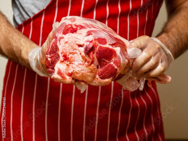 Fototapeta Fresh juicy leg of lamb with bone in a butcher hands. Red and white stripe apron background. Selective focus. High quality and price meat product. Agriculture and food industry.