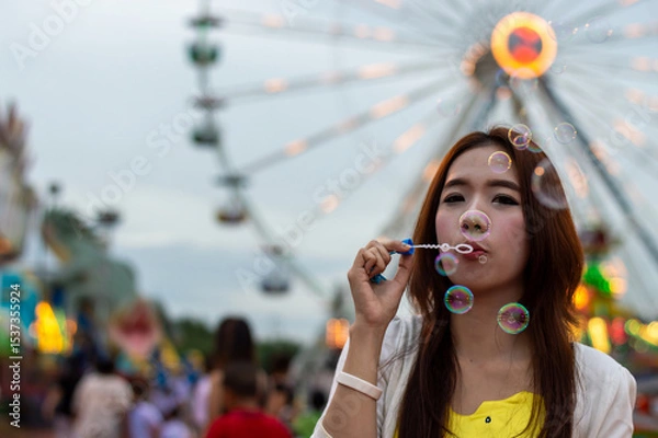 Obraz A young woman enjoying a day at the fair, blowing bubbles with a colorful ferris wheel in the background.