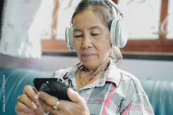 Obraz An elderly woman wears headphones while enjoying music on her smartphone, highlighting technology's impact on daily life.