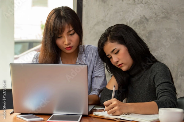 Obraz Two women collaborating on a project, one writing notes and the other working on a laptop in a modern workspace.