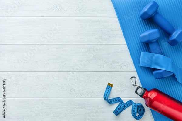 Obraz Fitness or healthy lifestyle concept. Blue dumbbells, red bottle, exercise mat and measuring tape on white wooden background with copy space. Minimal sports accessories idea. Top view with copy space