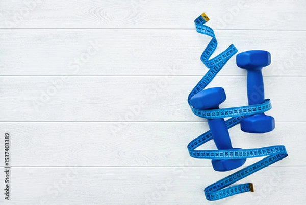 Obraz Fitness or healthy lifestyle concept. Blue dumbbells wrapped with blue measuring tape on white wooden background with copy space. Minimal sports accessories idea. Top view with copy space