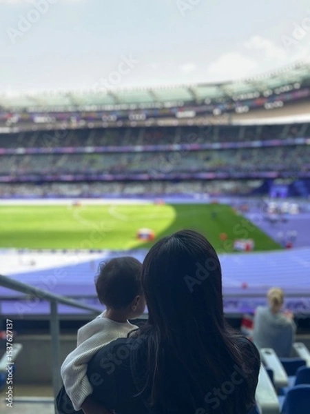 Fototapeta Behind view of a mother and her son enjoy watching sport at stadium. Quality time and generational sport supoorters in the crowd