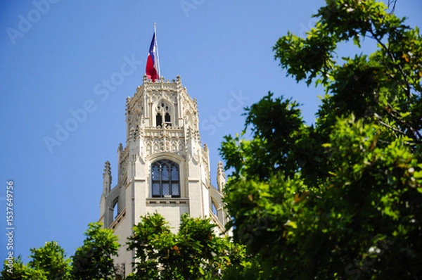 Fototapeta Historic tower with ornate architecture against blue sky and green foliage