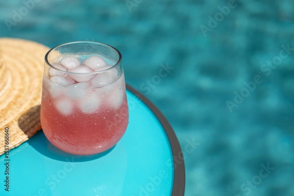 Obraz Refreshing pink beverage served in a glass filled with ice, on a turquoise table next to a straw hat, with a blurred blue swimming pool in the background with a space for text. summer vacation scene