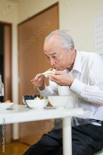 Fototapeta In a modest Japanese apartment, a man in his late 70s wearing a white shirt sits at a small table, eating a bowl of natto rice for breakfast as soft morning light filters gently through the window.