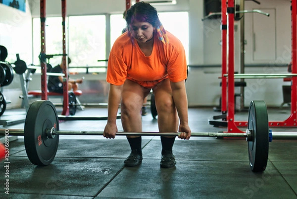 Fototapeta View of a beautiful, overweight young Latina lifting weights and working out at a gym. Positively, she's pushing herself to the limit. She's improving and feeling better.