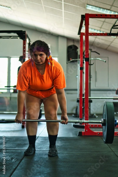 Fototapeta View of a beautiful, overweight young Latina lifting weights and working out at a gym. Positively, she's pushing herself to the limit. She's improving and feeling better.