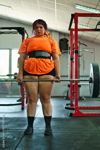 Fototapeta View of a beautiful, overweight young Latina lifting weights and working out at a gym. Positively, she's pushing herself to the limit. She's improving and feeling better.