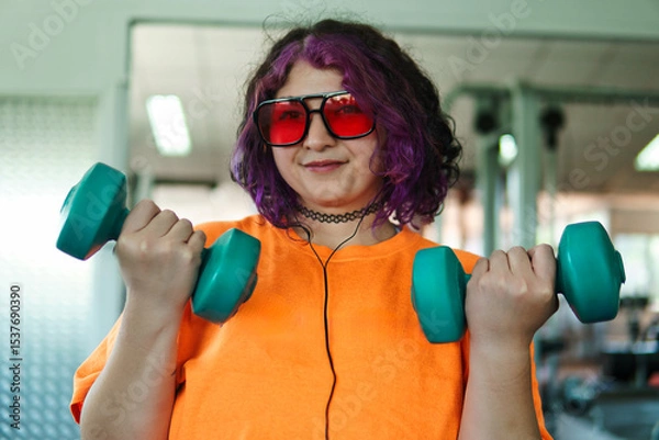 Fototapeta Beautiful young overweight Latina posing and exercising with weights and listening to music, wearing retro glasses and a Discman in a retro-style gym, working hard, very happy. Health and improvement.