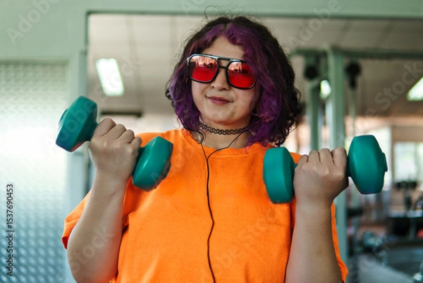 Fototapeta Beautiful young overweight Latina posing and exercising with weights and listening to music, wearing retro glasses and a Discman in a retro-style gym, working hard, very happy. Health and improvement.