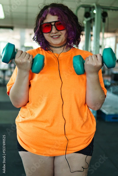 Fototapeta Beautiful young overweight Latina posing and exercising with weights and listening to music, wearing retro glasses and a Discman in a retro-style gym, working hard, very happy. Health and improvement.
