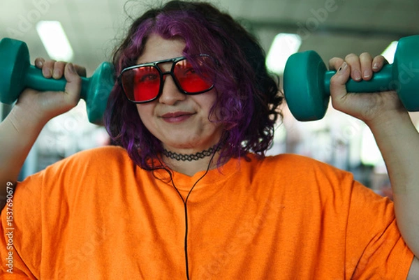 Fototapeta Beautiful young overweight Latina posing and exercising with weights and listening to music, wearing retro glasses and a Discman in a retro-style gym, working hard, very happy. Health and improvement.