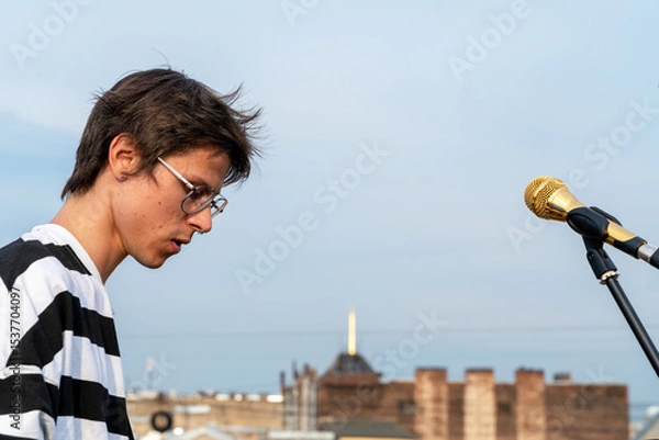 Fototapeta A young caucasian male in striped attire stands next to a microphone on a rooftop. the blurred urban backdrop and clear sky provide a vibrant setting, capturing creativity and urban life.