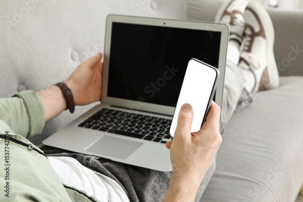 Fototapeta Handsome man with laptop using blank mobile phone on sofa at home, closeup