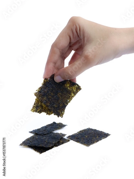 Fototapeta A hand picking up a square piece of dried seaweed snack with a stack of seaweed sheets on a white background, isolated for commercial food presentation or packaging use.