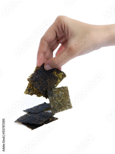 Fototapeta A hand picking up a square piece of dried seaweed snack with a stack of seaweed sheets on a white background, isolated for commercial food presentation or packaging use.
