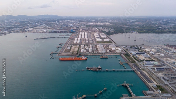 Obraz large oil tanker docked at a refinery terminal with multiple white storage tanks, complex pipeline networks, and offshore loading platforms. Represents global oil logistics