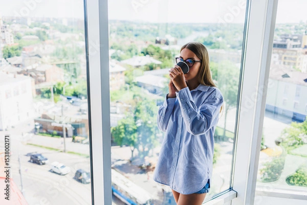 Fototapeta A young woman is savoring a cup of coffee while taking in the beautiful city view from her stylish, bright, and modern apartment. This scene reflects her relaxed lifestyle and peaceful outlook