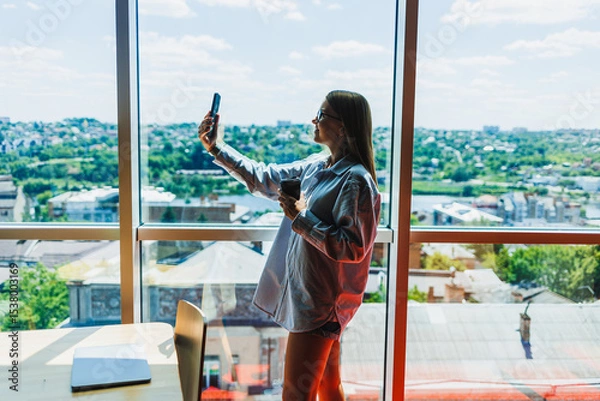 Fototapeta A young woman is happily taking a selfie with her smartphone in front of a scenic urban view seen through a large, modern window in a bright, airy space filled with daylight and warmth
