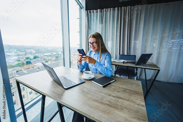 Fototapeta A dedicated businesswoman works remotely in her stylish office with a city view, using her smartphone to stay connected and productive, highlighting her commitment to innovative telecommuting