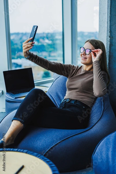 Fototapeta A young woman is savoring a peaceful moment as she takes a stylish selfie in a cozy environment, all while enjoying stunning views of the city. This upbeat moment captures happiness and leisure