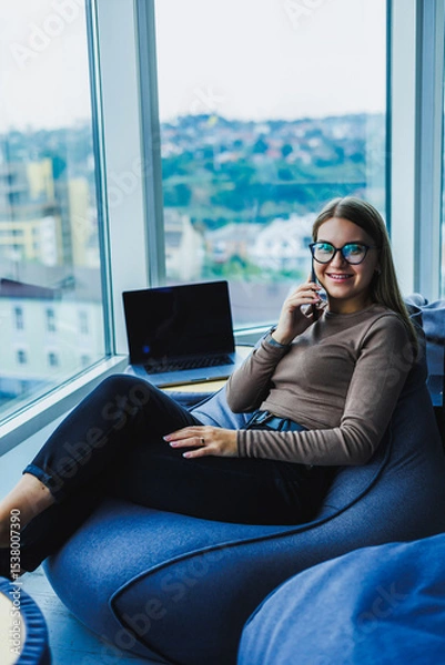 Fototapeta A dedicated young woman in a stylish outfit works hard in her beautifully decorated workspace, using modern technology and enjoying a stunning view of the vibrant cityscape through a large window