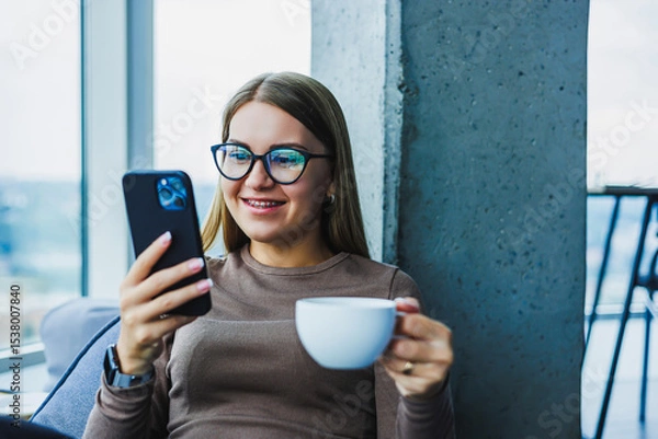 Fototapeta In a contemporary, stylish indoor environment, a young woman is fully immersed in enjoying her coffee while engaging with her smartphone, showcasing a perfect blend of leisure and modern lifestyle