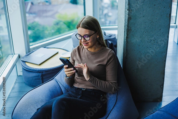 Fototapeta A young woman is happily engaging with her smartphone in a cozy and stylish workspace, embodying a sense of comfort and connectivity that reflects the latest modern lifestyle trends we see around us