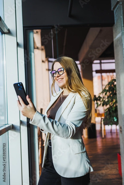 Fototapeta In a modern office, a confident businesswoman skillfully uses her smartphone, showcasing creativity and professionalism, embodying success and determination, and inspiring others around her