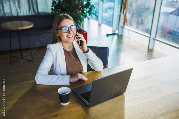 Fototapeta In a modern office, a professional woman is on a crucial business call, her laptop open and a fresh cup of coffee by her side, showcasing her focus and productivity during work hours