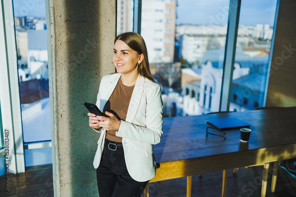 Fototapeta A confident businesswoman is engaged with her smartphone in a modern office setting, showcasing her proficient skills in entrepreneurship and networking through digital means and technology