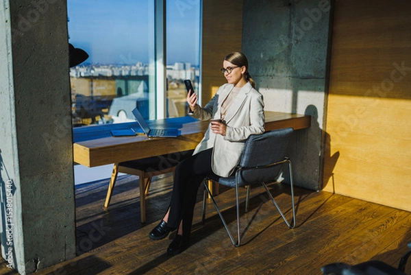 Fototapeta A chic and stylish businesswoman is enjoying her coffee while engaging in a phone interaction within a contemporary modern office setting that reflects elegance and professionalism