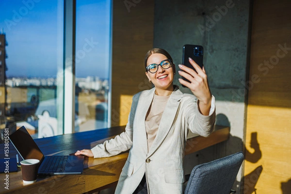 Fototapeta A confident and professional woman happily taking a selfie in a modern office setting that boasts a stunning city view, reflecting her successful career and trendy lifestyle in the business world