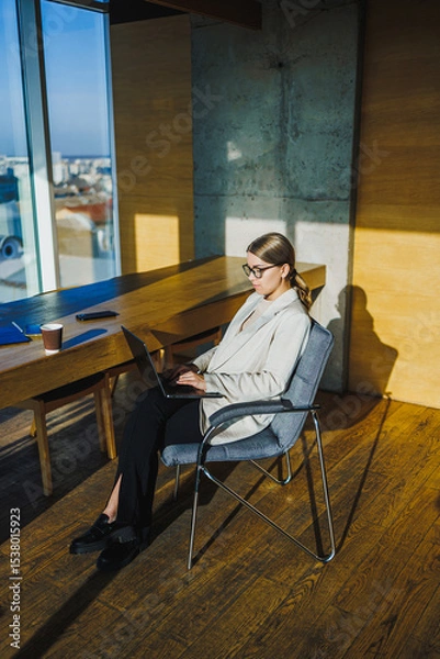 Fototapeta A dedicated professional woman engages in her work remotely from a contemporary office environment, featuring expansive windows that allow plenty of natural light to illuminate the space