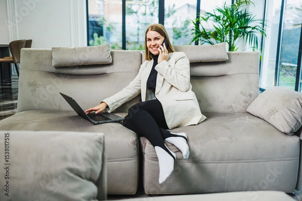 Fototapeta A professional woman is working from home on her laptop, engaged in conversation while sitting comfortably on a cozy sofa in her modern living space, blending productivity with relaxation