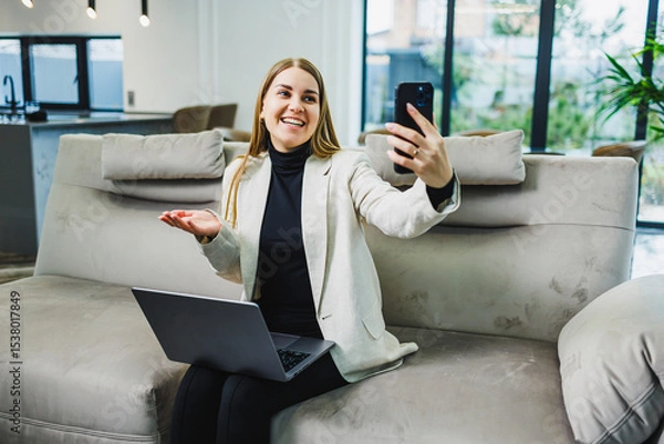 Fototapeta A professional businesswoman is actively engaged in a video call while comfortably seated on her couch, using her laptop to facilitate effective communication and collaboration with team members