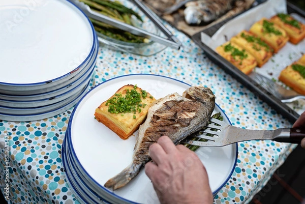 Fototapeta Grilled fish with gratin and chives on plate. Preparation food on a table for lunch or dinner on barbeque. Healthy mediterranean meal during garden party or in restaurant. Dorada fish ready to eat.