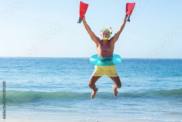 Obraz Senior man jumping midair on sandy shore, wearing snorkel mask with green ring and red fins