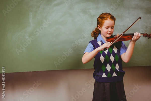 Fototapeta Schoolgirl playing violin with bow before green chalkboard in classroom wearing uniform, copy space