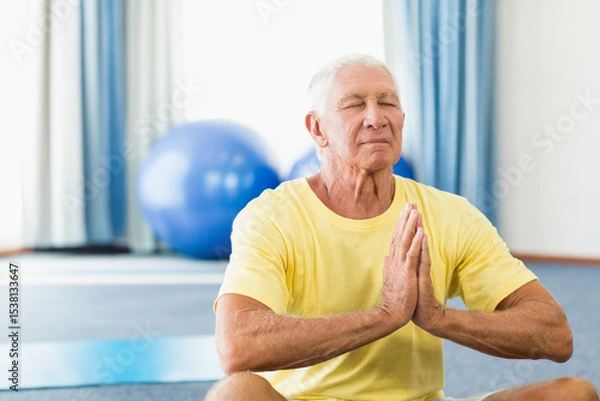 Fototapeta Senior man meditating on yoga mat in gym with stability balls while wearing yellow t-shirt