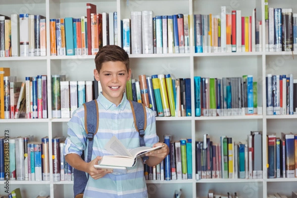Fototapeta School-age boy standing in school library wearing backpack reading open book before white bookshelf
