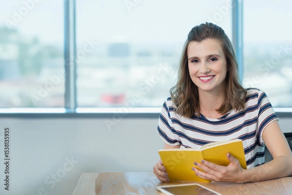 Fototapeta Woman reading yellow paperback book at wooden table near city window with tablet nearby, copy space